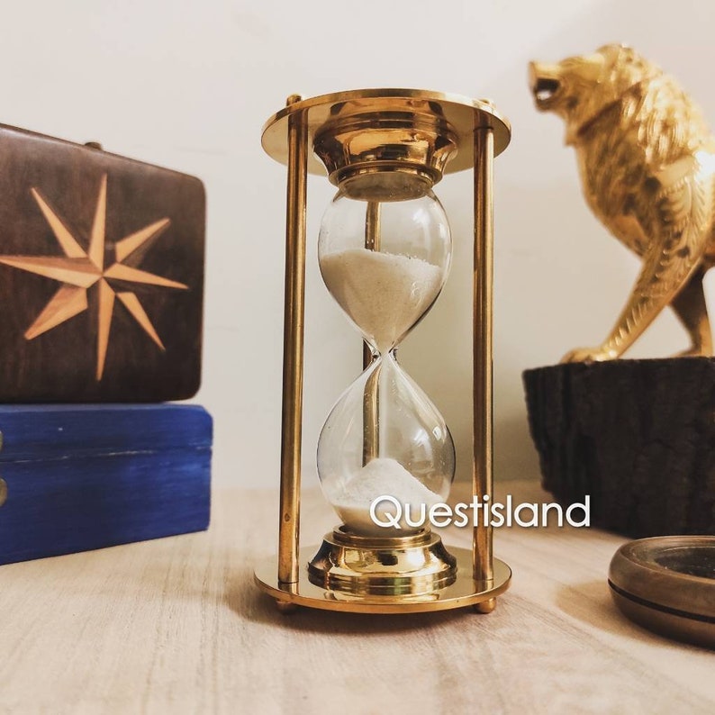 May include: A brass hourglass with white sand, standing on a wooden surface. The hourglass is about 6 inches tall and has a classic design.