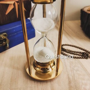 May include: A gold hourglass with two glass bulbs filled with white sand. The hourglass is on a wooden surface.