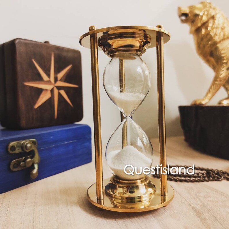 May include: A brass hourglass with white sand, sitting on a wooden surface. The hourglass is about 6 inches tall and has a clear glass bulb. The sand is flowing from the top bulb to the bottom bulb. The hourglass is a decorative item and is perfect for adding a touch of elegance to any room.