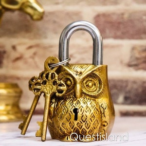 Puede incluir: Un candado dorado en forma de búho con dos llaves. El candado tiene un diseño de búho detallado con un patrón de diamante en su cuerpo. Las llaves son ornamentadas y tienen un aspecto vintage.