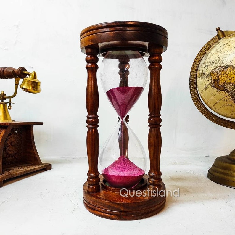 May include: A wooden hourglass with a dark brown finish and a pink hourglass sand. The hourglass is on a white surface.