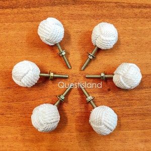 May include: Six white rope knob pulls with gold screws. The knobs are arranged in a circle on a wood surface.