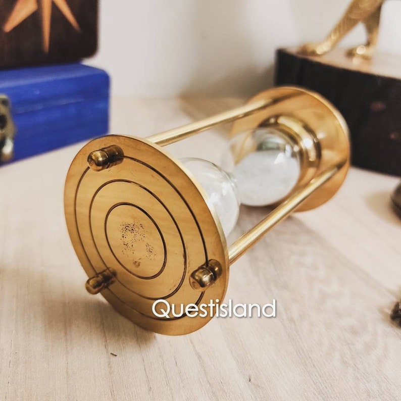 May include: A gold hourglass with a round base and a clear glass bulb filled with white sand. The hourglass is on a wooden surface.