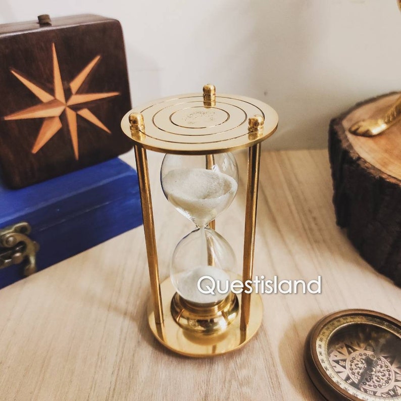 May include: A gold-toned hourglass with a clear glass bulb. The hourglass is sitting on a wooden surface. The sand is white.