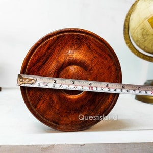 May include: A close-up of the bottom of a wooden pedestal with a brown finish. A tape measure is placed across the bottom of the pedestal, showing a measurement of 10 inches.