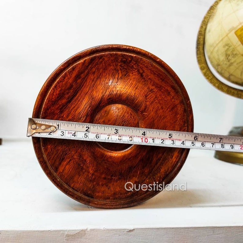 May include: A close-up of the bottom of a wooden pedestal with a brown finish. A tape measure is placed across the bottom of the pedestal, showing a measurement of 10 inches.