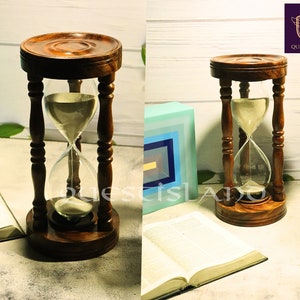 May include: A wooden hourglass with a clear glass bulb filled with sand. The hourglass is sitting on a book with a blue and yellow box behind it. The hourglass is made of dark wood and has a round base.