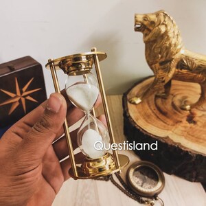 May include: A gold hourglass with white sand, sitting on a wooden surface. The hourglass is in a hand, and there is a gold lion statue and a compass in the background.