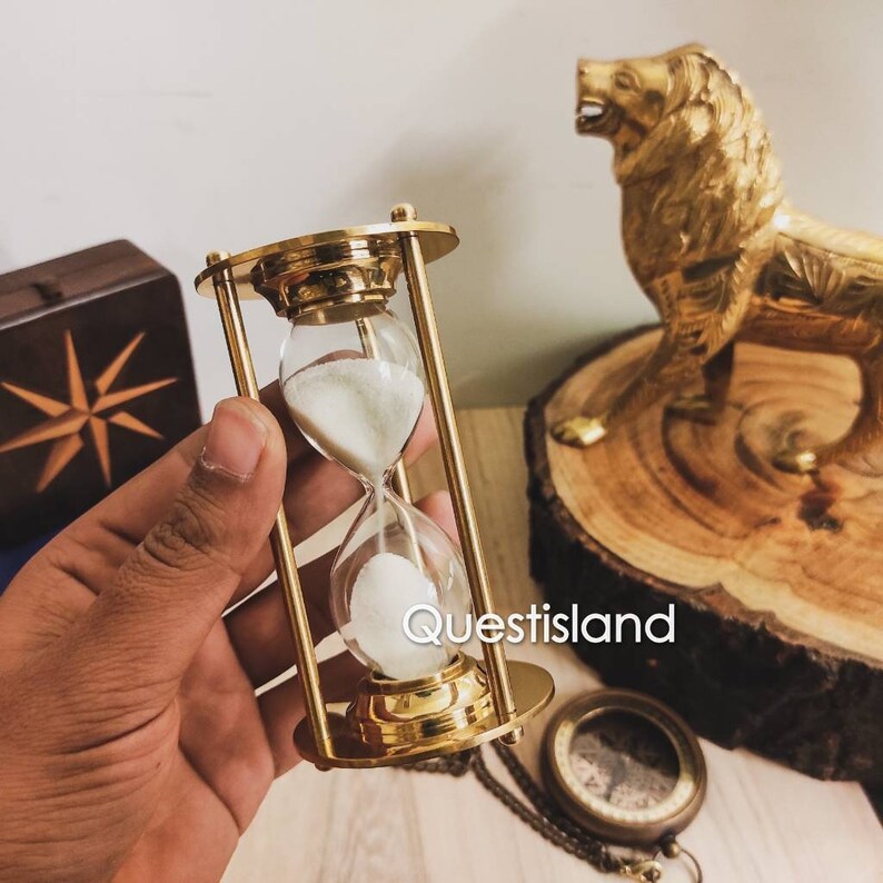 May include: A gold hourglass with white sand, sitting on a wooden surface. The hourglass is in a hand, and there is a gold lion statue and a compass in the background.