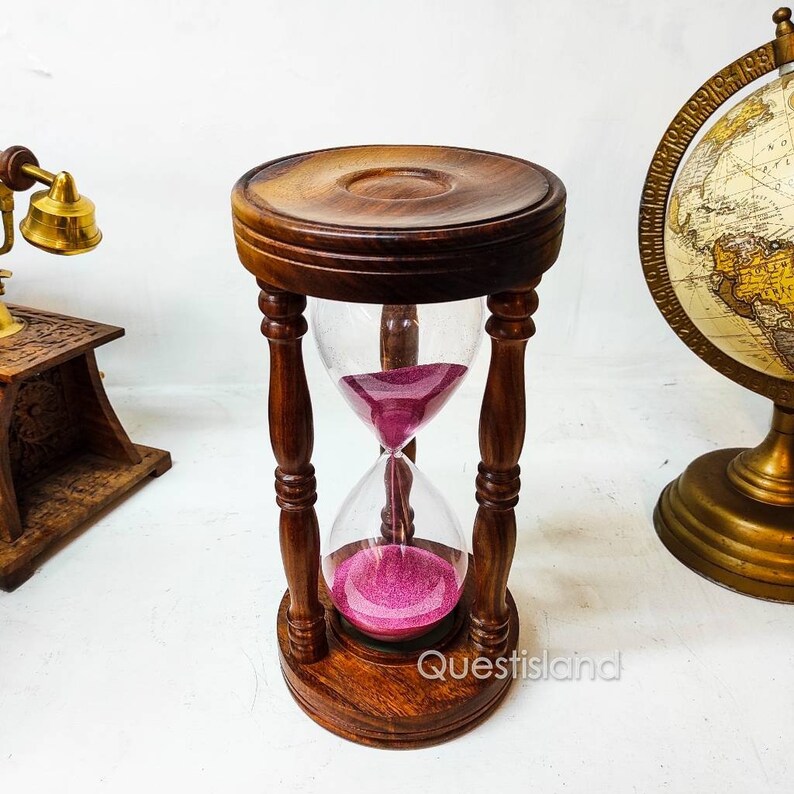 May include: A wooden hourglass with a pink sand timer. The hourglass is made of dark wood and has a glass bulb. The hourglass is sitting on a white surface.