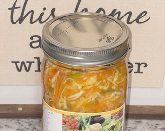 May include: A glass jar filled with a yellow pickled vegetable mixture. The jar has a yellow label with the text "Pik-nix" and "manchma delicious" printed on it. The jar is sitting on a light gray countertop.