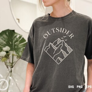 May include: A dark gray t-shirt with a white graphic design of mountains and the word "OUTSIDER" in a curved font. The graphic is in a diamond shape.