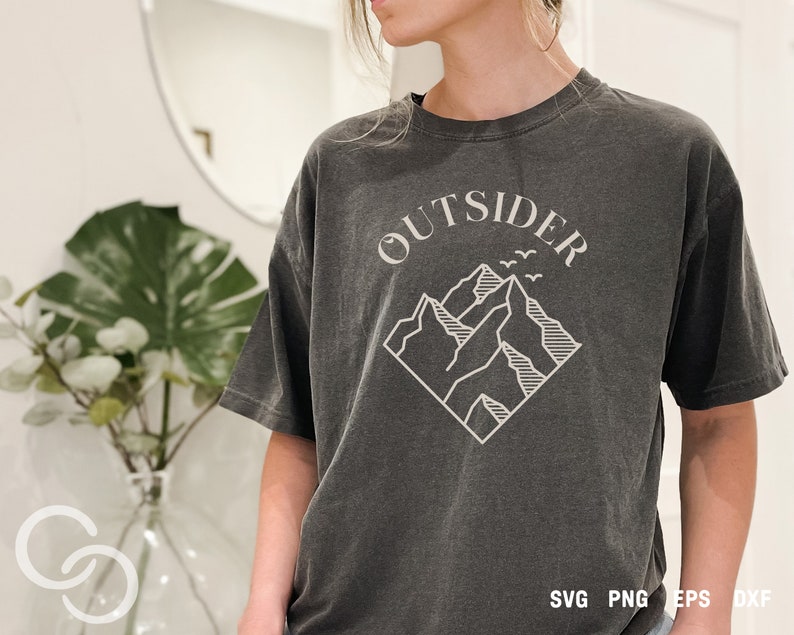 May include: A dark gray t-shirt with a white graphic design of mountains and the word "OUTSIDER" in a curved font. The graphic is in a diamond shape.