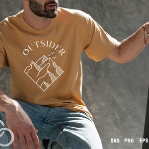 May include: A person wearing a mustard yellow t-shirt with a white graphic of mountains and the word "OUTSIDER" above the mountains. The person is holding a skateboard.