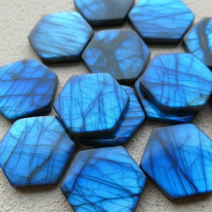 Puede incluir: Primer plano de varias piedras hexagonales de labradorita pulida. Las piedras son de un azul vibrante con patrones intrincados en forma de telaraña y vetas de color gris oscuro. Ideales para la creación de joyas.