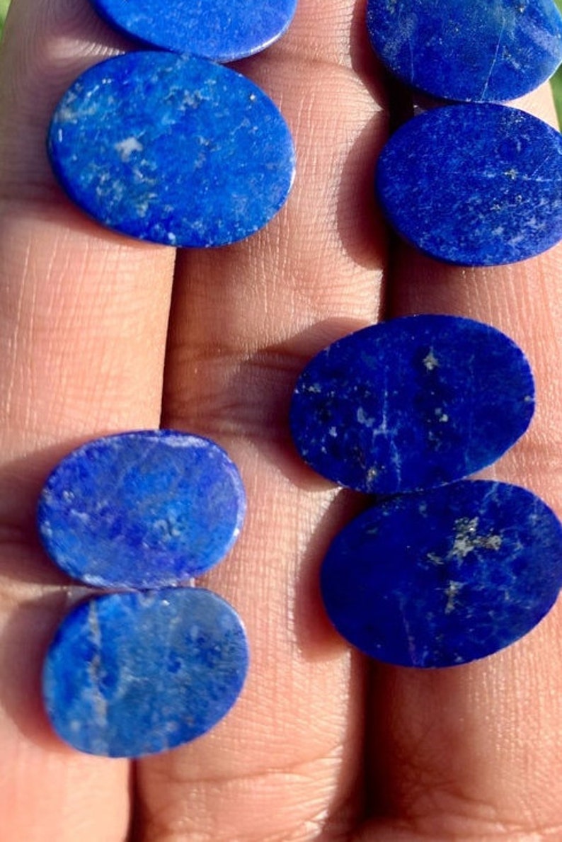 Op de afbeelding: Een verzameling ovale lapis lazuli cabochons, vari&euml;rend in grootte, met een diepblauwe kleur en witte adering. De stenen zijn glad en gepolijst, met een natuurlijke, aardse uitstraling.