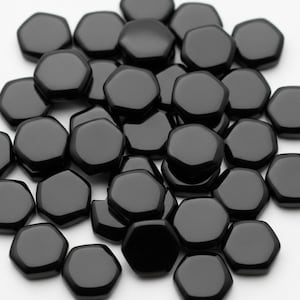 以下が含まれることがあります： 小さな黒色の六角形のビーズのコレクション。ビーズは滑らかで光沢のある表面を持ち、白い背景に散らばっています。形状は均一で、全体的な効果はシンプルなエレガンスです。