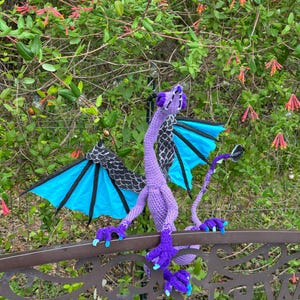 Puede incluir: Una escultura de dragón de ganchillo hecha a mano en tonos morado, negro y azul. El dragón tiene alas extendidas, escamas detalladas y está posado en una barandilla de metal decorativa. El fondo presenta follaje verde y flores rojas.