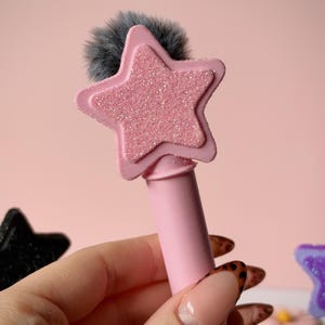 以下が含まれることがあります： 淡いピンク色の円筒形ツールで、上部には質感のあるキラキラしたピンク色の星型があしらわれています。星型の後ろには柔らかいダークグレーのフェイクファーのポンポンが付いており、星の上端から少しだけはみ出しています。