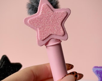 Puede incluir: Una herramienta cilíndrica de color rosa claro con una parte superior en forma de estrella que presenta una superficie rosa texturizada y brillante. Un pompón de piel sintética gris oscuro está sujeto detrás de la estrella, sobresaliendo ligeramente.