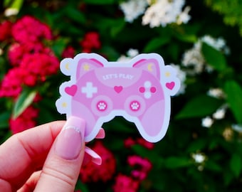 Könnte beinhalten: Rosa-weißer Controller-Aufkleber mit Katzenohren und Pfotenabdrücken. Der Aufkleber lautet "Let's Play".