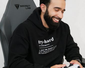 May include: A person wearing a black hoodie with the text "try-hard" and a definition of the word. The person is sitting in a black and gray gaming chair and holding a white video game controller.