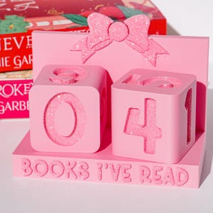 May include: Pink wooden block calendar with glitter accents, displaying the numbers "04". The base reads "BOOKS I'VE READ". A pink bow is at the top. Books are in the background.