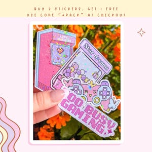 May include: Four pink and purple holographic stickers with glitter. The stickers feature a video game controller, a retro arcade game, a star, and the text "Do Busy Gaming".