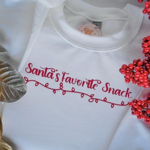 Peut inclure: Un body blanc pour bébé avec l'inscription rouge "Santa's Favorite Snack". Le texte est souligné par une guirlande lumineuse rouge. Décoré de baies rouges et de feuilles dorées et argentées.