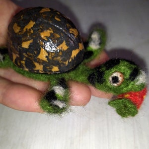 Puede incluir: Una pequeña tortuga de fieltro hecha a mano con un caparazón marrón y amarillo, un cuerpo verde y ojos negros. La tortuga sostiene una flor roja de fieltro en su boca.