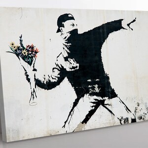 May include: Black and white stencil art print of a person throwing a bouquet of colorful flowers. The person is wearing a black hooded sweatshirt and a black mask. The background is a white wall with some texture.