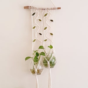 May include: A white macrame plant hanger with two glass globes, each containing a green plant. The hanger is decorated with green felt leaves and has a wooden dowel at the top.