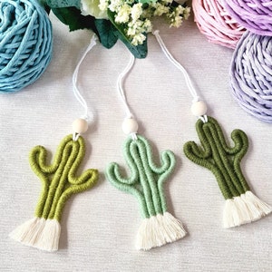 May include: Three green and white macrame cactus ornaments with wooden beads and white tassels. The ornaments are hanging from white cords.