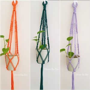 May include: Three macrame plant hangers with a small potted plant in each. The hangers are made with thick cord and have a tassel at the bottom. The hangers are orange, green, and purple.