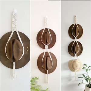 May include: Three brown hats hanging on a white macrame wall hanging. The macrame is made with white cord and has a knotted design. The hats are all different sizes and shapes.