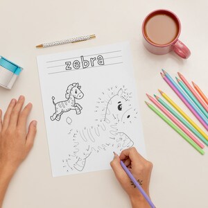 K&ouml;nnte beinhalten: Ein Kind verbindet die Punkte auf einer Schwarzwei&szlig;zeichnung eines Zebras auf einem wei&szlig;en Blatt Papier. Das Wort "Zebra" steht oben auf der Seite. Das Kind verwendet einen violetten Stift, um die Punkte zu verbinden. Auf dem Tisch liegen verschiedene farbige Stifte.