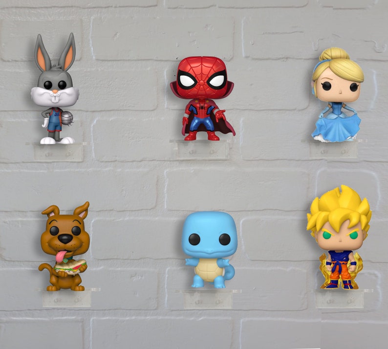 Op de afbeelding: Een verzameling van zes pop vinyl figuren die op een witte bakstenen muur worden getoond. De figuren omvatten Bugs Bunny in een basketbaluniform, Spiderman in zijn rood-blauwe pak, Assepoester in een blauwe jurk, Scooby Doo met een sandwich, een blauw dinosaurusachtig wezen en een blond anime-personage in een rood-oranje outfit.
