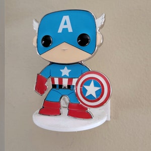 Op de afbeelding: Een klein, zilveren 3D-figuurtje van Captain America. Het figuurtje draagt een blauw en rood kostuum met een witte ster op de borst. Het figuurtje houdt een rood, wit en blauw schild vast met een witte ster in het midden. Het figuurtje staat op een witte basis.