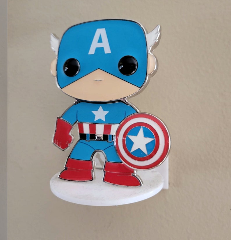 Op de afbeelding: Een klein, zilveren 3D-figuurtje van Captain America. Het figuurtje draagt een blauw en rood kostuum met een witte ster op de borst. Het figuurtje houdt een rood, wit en blauw schild vast met een witte ster in het midden. Het figuurtje staat op een witte basis.