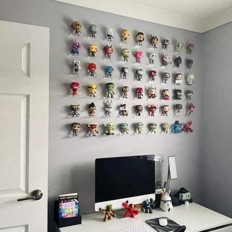 May include: A wall display of various colorful Funko Pop figures on clear acrylic shelves. The figures are arranged in rows and columns, with a variety of characters and designs. The wall is painted a light gray color, and there is a desk with a computer monitor and other items in the foreground.