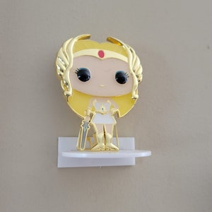 May include: Gold-colored She-Ra Pop figure with a white dress and a sword. The figure is on a white shelf.