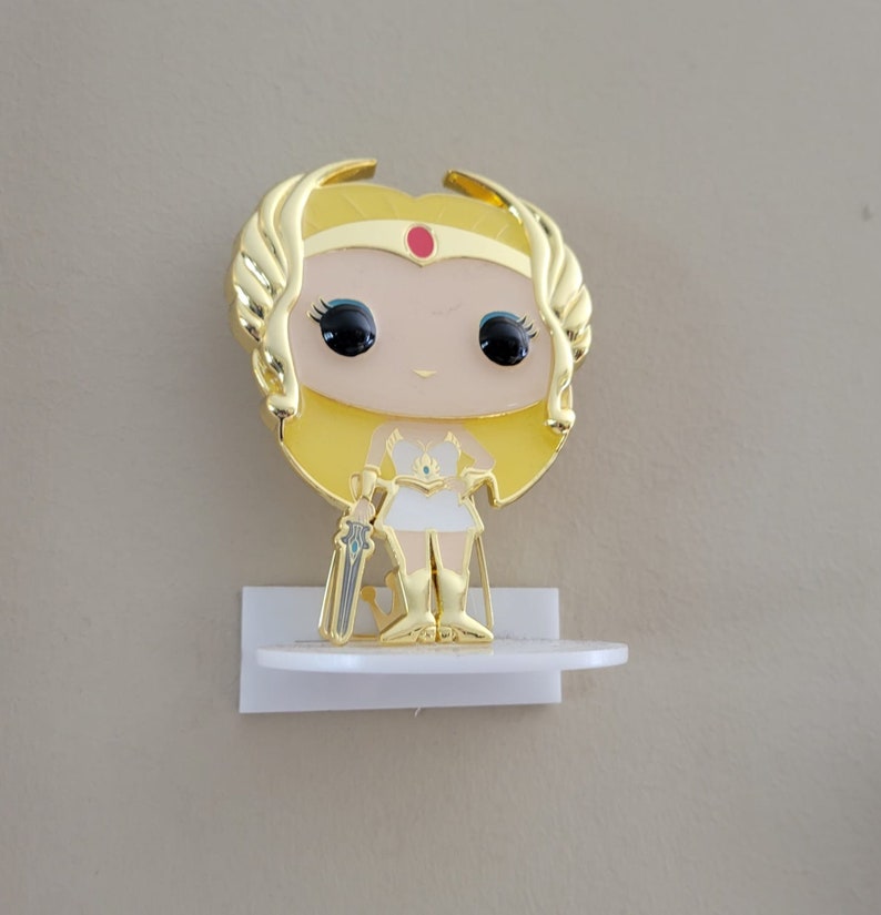 May include: Gold-colored She-Ra Pop figure with a white dress and a sword. The figure is on a white shelf.