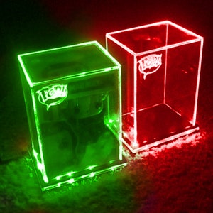 May include: Two clear acrylic display cases, one illuminated with green light and the other with red. Each case has a "POP!" logo. The green case has a figure inside. The cases are designed to showcase collectibles.