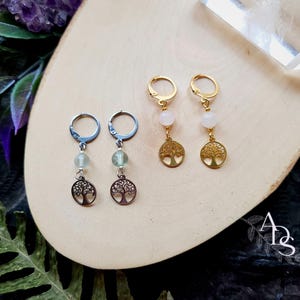 Peut inclure: Boucles d'oreilles avec un motif d'arbre de vie. Deux paires sont présentées. L'une a des créoles argentées, des perles vertes et des pendentifs en forme d'arbre. L'autre a des créoles dorées, des perles blanches et des pendentifs en forme d'arbre.