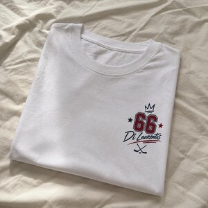 May include: A white t-shirt featuring a graphic on the lower right side. The design includes the number 66 in red with a dark outline, a small crown icon above it, two stars on the sides, the text Di Laurentis, and two crossed hockey sticks.