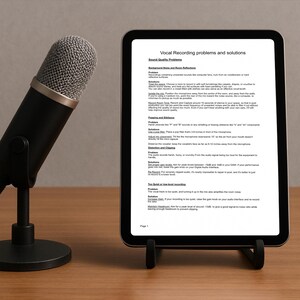 Puede incluir: Un micrófono negro con una cabeza de malla plateada se encuentra a la izquierda de una tableta que muestra texto sobre problemas y soluciones de grabación vocal. La tableta está sobre un soporte negro en una superficie de madera.