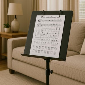 Puede incluir: Un atril negro con partituras que muestra el texto "Sheet Music Cheatsheet". El atril es ajustable y tiene un pequeño estante. El fondo incluye un sofá, una lámpara y una mesa auxiliar.