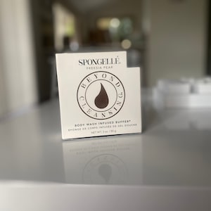 Pode incluir: Caixa branca com o texto "SPONGELLÉ FREESIA PEAR BEYOND CLEANSING" e uma gota de líquido marrom em um círculo. O texto "BODY WASH INFUSED BUFFER* EPONGE DE CORPS INFUSÉE DE GEL DOUCHE NET WT. 3 oz/85g" está impresso na parte inferior da caixa.