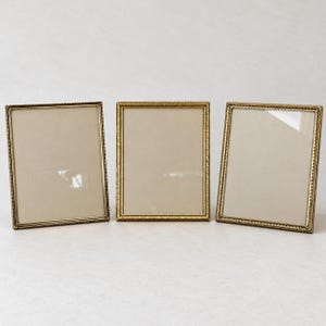 May include: Three rectangular picture frames with a metallic gold finish and a textured, beaded edge detail. Each frame features a clear glass front and a neutral-colored backing, standing upright in a row against a plain, light-colored background.