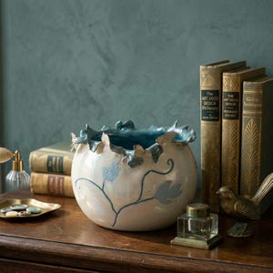 May include: A ceramic bowl with a ruffled rim and a pearlescent finish. The bowl features a blue interior and is decorated with blue leaf and vine designs. The bowl is displayed on a wooden surface with books and decorative items.